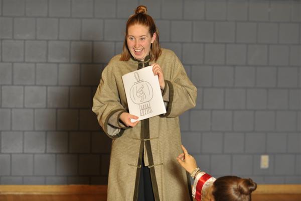 A person smiling and holding a sketch on paper, showing it to someone partially visible in the foreground. The person is wearing a light-colored robe and standing against a background of gray tiles.