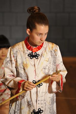 Person wearing a patterned robe with black knot buttons holds a bamboo stick, looking focused. Hair is in a bun, and the background includes another person partially visible. The setting appears to be indoors, with dim lighting and a grey wall.