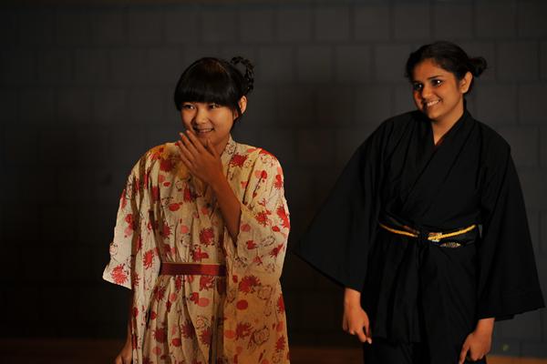 Two individuals stand together, smiling. One wears a floral-patterned kimono with red accents, and the other wears a dark robe with a gold belt. The background is a dimly lit wall.