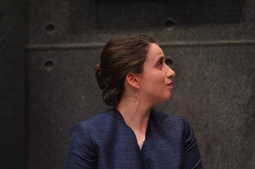 Woman with dark hair in a bun, wearing a navy blazer, looking to her left against a textured gray wall with circular details.