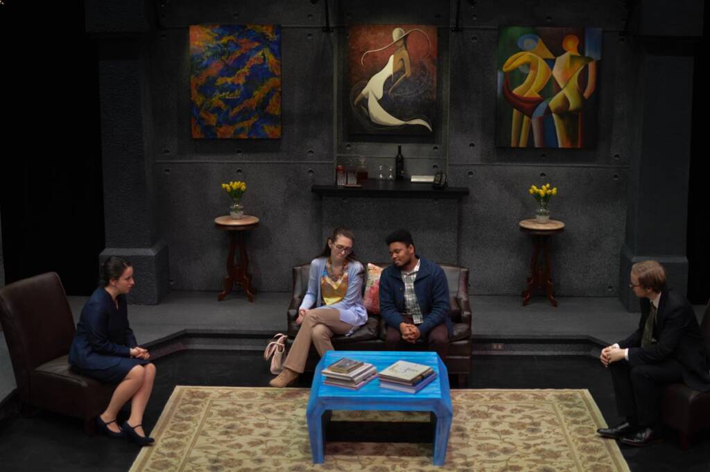 A group of four people sits in a living room decorated with abstract paintings. Two vases with yellow flowers are on tables flanking a fireplace. Stacks of books are on a blue coffee table in the center. The mood is contemplative.