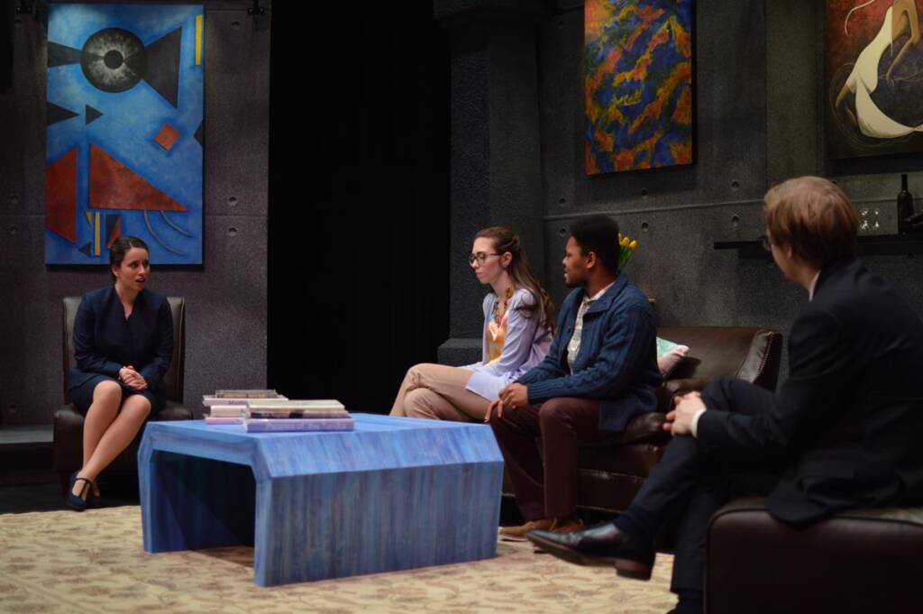 Four people are seated in a living room setting engaged in conversation. The room has abstract artworks on the walls and a blue coffee table with books. The atmosphere appears formal and attentive.