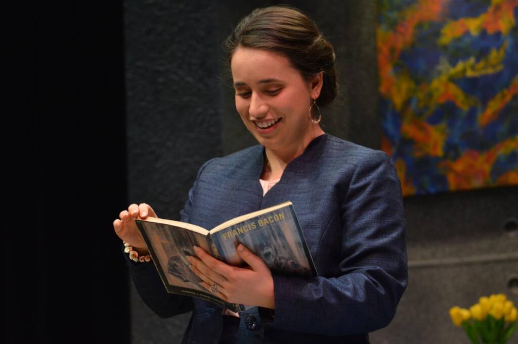 A person with brown hair, wearing a navy blazer, is smiling while reading a book titled "Francis Bacon." They are indoors, with a colorful painting and yellow flowers visible in the background.