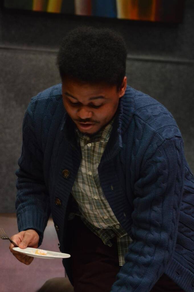 A person wearing a blue sweater and checkered shirt is leaning forward, holding a paper plate with food and a fork. The background is blurred, with dark gray walls and a colorful abstract painting.