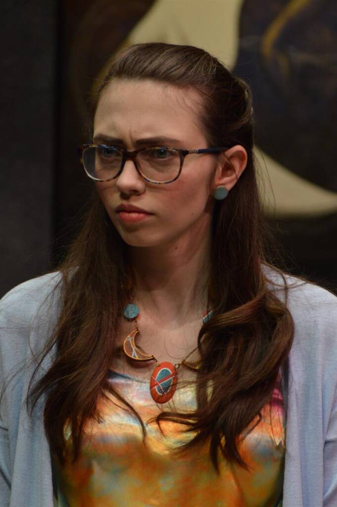 A person wearing glasses looks serious while standing indoors. They have long brown hair, and are dressed in a colorful top with a statement necklace and blue earrings. The background is dark and out of focus.