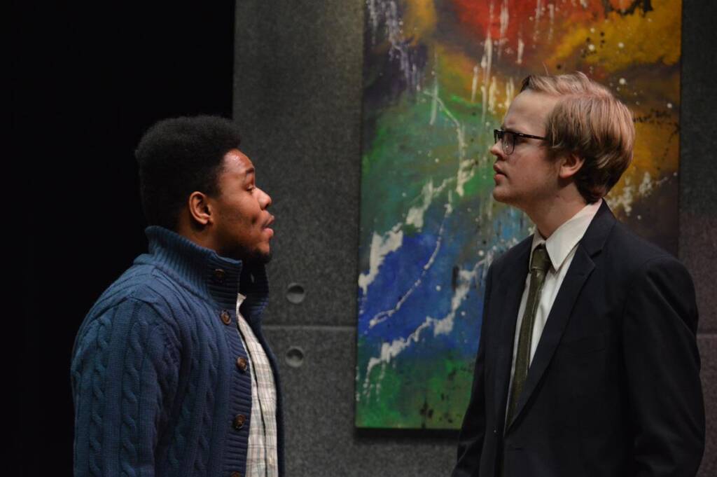 Two actors perform on stage. One wears a blue sweater over a checked shirt, and the other wears a suit and glasses. A colorful abstract painting is in the background.