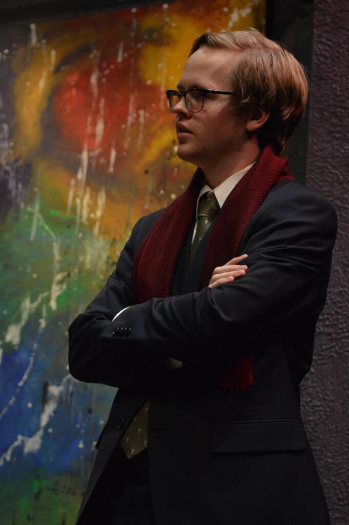 A man with glasses and light brown hair stands with arms crossed, wearing a black suit, green tie, and dark red scarf. He is in front of a colorful abstract painting with splashes of red, yellow, green, and blue.