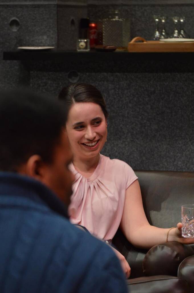 A woman in a light pink blouse sits on a sofa, holding a glass. She is smiling and looking at a person in the foreground who is wearing a dark jacket. The background includes a shelf with various items.