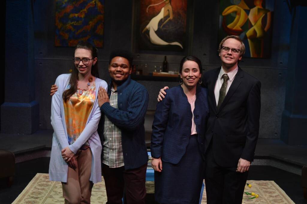 Four people stand on a stage, posing together. The background features abstract paintings. They are dressed formally; one wears a tie. The group appears cheerful, with some having their arms around each other.