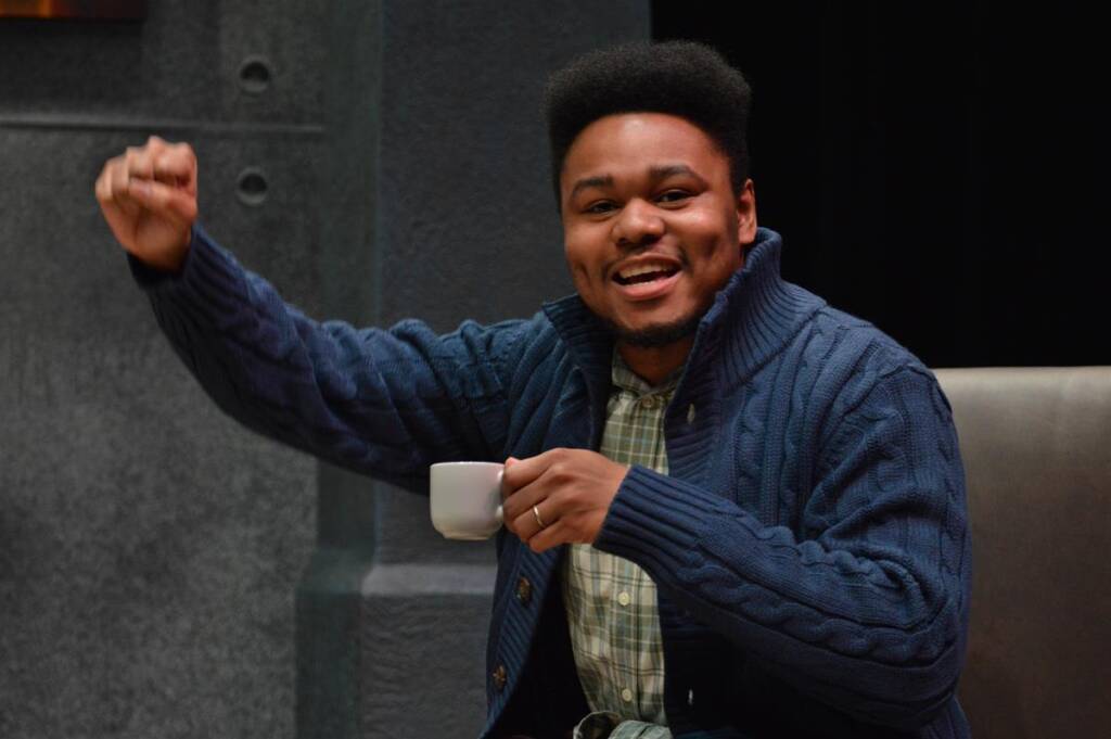 A person in a blue sweater and checkered shirt holds a small white cup. They are smiling and raising their other hand as if gesturing enthusiastically, sitting against a dark backdrop.