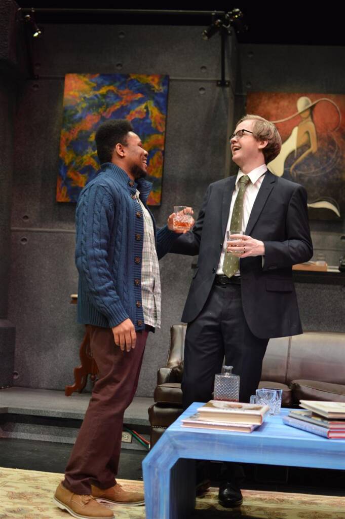 Two men are laughing and clinking glasses in a room with abstract paintings on the wall. One man wears a blue sweater, and the other is in a suit and tie. A stack of books and a crystal decanter are on the table in front of them.