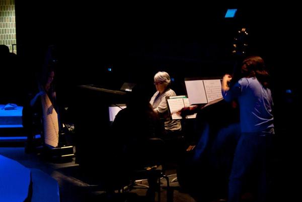 A dimly lit music performance setup with musicians focused on sheet music. A woman with short white hair sits at a piano or keyboard, and a person with long hair nearby plays a string instrument. Blue lighting casts a subtle glow on the scene.