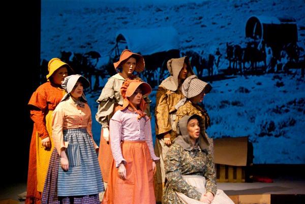 A group of people in pioneer attire, including bonnets and aprons, perform on stage. Behind them, a backdrop shows a wagon train scene. Their expressions suggest they're singing or speaking as part of a theatrical production.