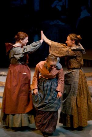 Three people in period costumes stand on stage. Two of them are giving a high-five to each other, while the third person in the middle is slightly bent forward, smiling. The background is dark, suggesting a theater setting.