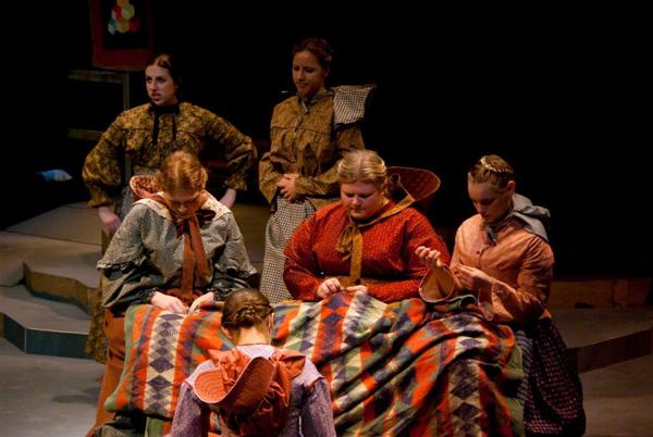 A group of people in period costumes work together on a colorful, patterned quilt. They are focused on sewing, with one person standing in the background. The scene appears to be part of a theatrical performance.