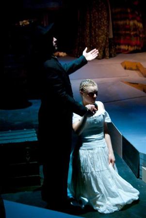 A man in dark clothing stands with one arm extended, holding hands with a woman in a white dress. She is kneeling on a dimly lit stage, creating a dramatic atmosphere.