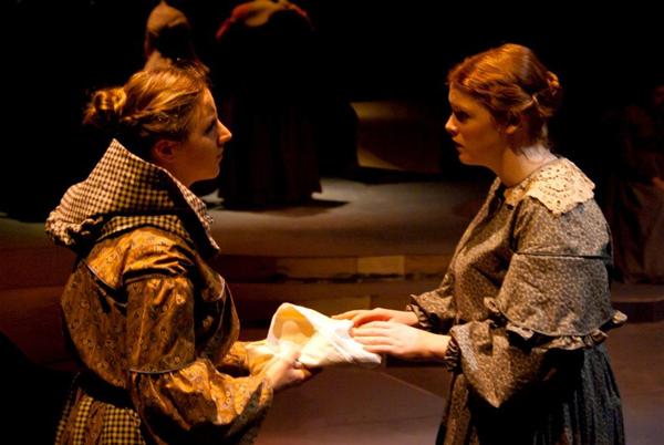 Two women in period costumes engage intensely on a dimly lit stage, holding a white cloth between them. The scene is serious, suggesting a dramatic moment in a historical play.