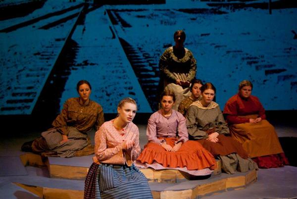 Seven women in period clothing sit on a stage with a backdrop of train tracks. One woman is seated at the front, appearing to speak or perform. The scene suggests a theatrical performance or play.