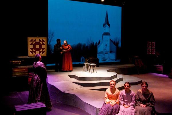 A theater stage set with a rural scene backdrop showing a church. Actors are dressed in period costumes; one stands with others seated in the foreground, observing. The lighting sets a serene, reflective mood.