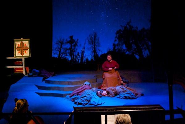 A stage scene depicts a person sitting on stone steps against a starry night backdrop. Several figures are lying nearby, covered in blankets. A quilt with a geometric design hangs on the left. A person is partially visible at the bottom.