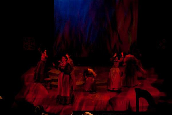 A dimly lit stage with performers in flowing costumes, appearing to be engaged in a dynamic dance or theatrical performance. The scene is filled with red and blue lighting, creating a dramatic atmosphere.