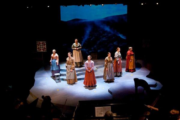 Seven people in period costumes stand on a dimly lit stage with a backdrop featuring a mountainous landscape. The stage has a simple set, and a piano is visible in the foreground. They seem to be in a theatrical performance.