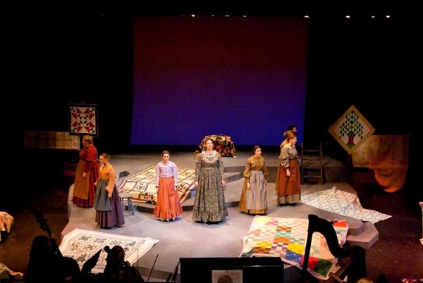 A theater stage featuring seven performers in period costumes. Quilts and patchwork designs are displayed around them. The background is a plain, illuminated screen. An orchestra pit with musicians is visible in the foreground.