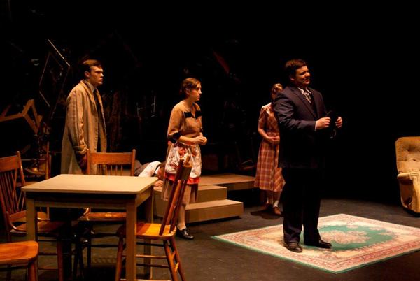 Four people in a dimly lit theater stage setting. A table with chairs and a rug are in the foreground. One person is dressed in a suit, while the others wear casual attire, standing and sitting in a living room-like arrangement.