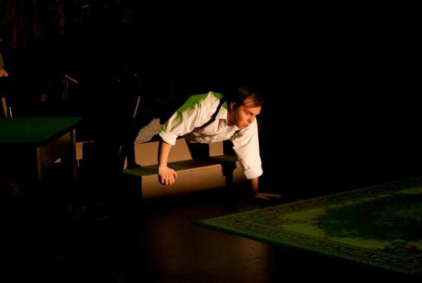 A person in a white shirt and dark pants is leaning forward on a set of steps, reaching toward a green patterned rug. The scene is dimly lit, creating a dramatic atmosphere.