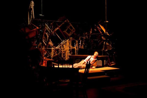 A person lies on a dimly lit stage, surrounded by an arrangement of chairs and ropes hanging above. The scene is dramatic and cluttered, with a dark, mysterious atmosphere.