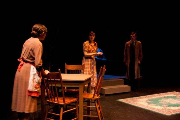 Three people on a dimly lit stage perform a scene. One person stands by a table with chairs, wearing an apron. Another, in a patterned dress, gestures while speaking. The third person, in a long coat, stands to the side. A patterned rug is visible.