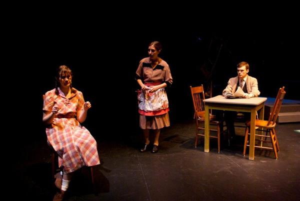 Three people are on stage in a dimly lit theater. A woman sits on the left wearing a plaid dress, gesturing. Another woman stands in the middle wearing an apron. A man sits at a table on the right, looking towards the woman on the left.