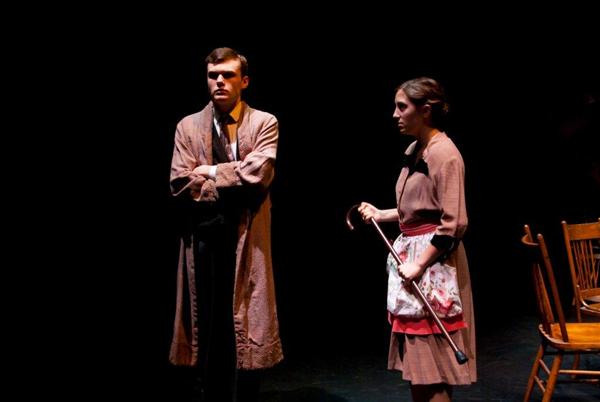 A man and woman in period costumes perform on stage under dim lighting. The man stands with arms crossed, while the woman holds a cane and wears an apron. Wooden chairs are visible in the background.