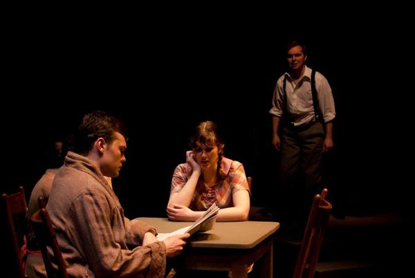 Three people are on stage in a dramatic play. One sits at a table reading a newspaper, while another rests her head on her hand looking contemplative. A third person stands in the background, wearing a shirt with suspenders, creating a tense atmosphere.