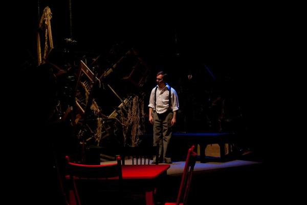 A person stands on a dimly lit stage, dressed in a white shirt and suspenders. The background is a chaotic arrangement of chairs and ropes. A red chair and table are visible in the foreground.