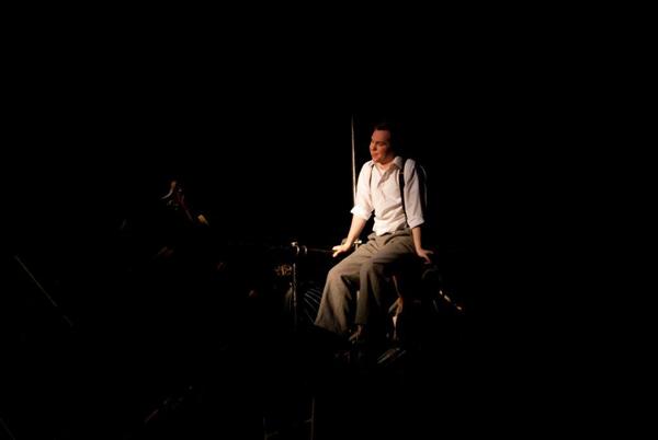 A person in a white shirt and suspenders sits on a dimly lit stage, surrounded by darkness, creating a dramatic and introspective atmosphere.