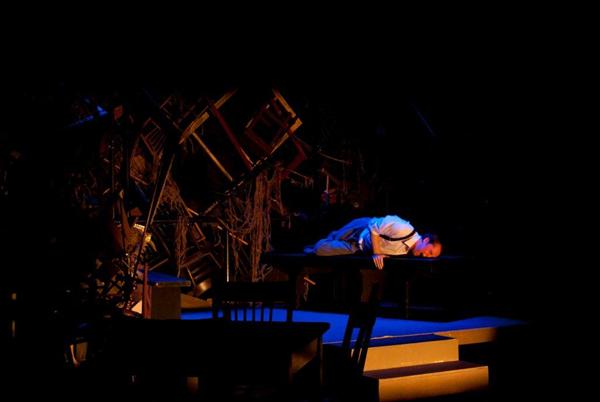 A person in a white shirt and suspenders lies face down on a table, surrounded by a dimly lit stage with stacked chairs and wooden debris in the background. The scene is dark with dramatic lighting highlighting the figure.