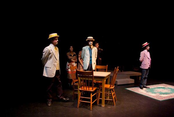 Five people in colorful period costumes stand on a dimly lit stage. A wooden table with chairs is in the center. One person stands on a rug. The backdrop is dark, highlighting the performers.