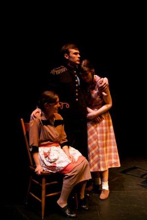 A theatrical scene with a person in a military-style uniform standing, comforting two seated women in vintage dresses on a dimly lit stage. One woman leans on the standing person, while the other sits in a chair.