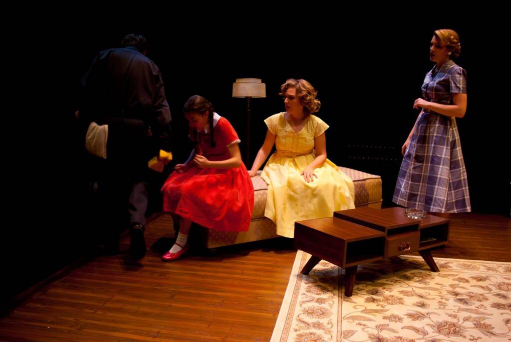 Four people perform on a dimly lit stage with retro furniture. Three women in vintage dresses sit on a couch, two in conversation and one looking away. A person in dark clothing walks off stage, adding movement to the scene.