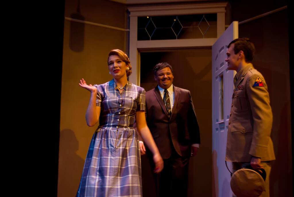 Three people stand in a warmly lit room. A woman in a plaid dress is gesturing and smiling. Behind her, two men smile; one in a suit, the other in a military uniform holding a hat. An open door is in the background.