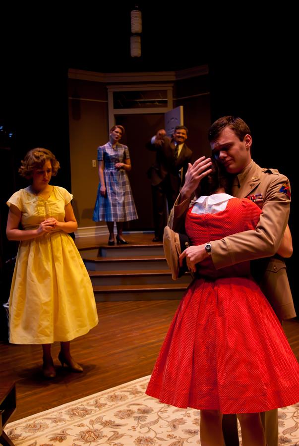 A stage scene with a man in a beige uniform hugging a woman in a red dress. A woman in a yellow dress stands nearby, looking pensive. In the background, two people are on stairs, observing the scene.