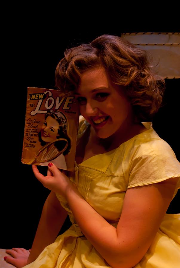 A woman in a yellow dress smiles while holding up a vintage magazine titled "New Love." The cover features a photo of a smiling woman. The background is dark, highlighting the woman's joyful expression and the retro magazine.