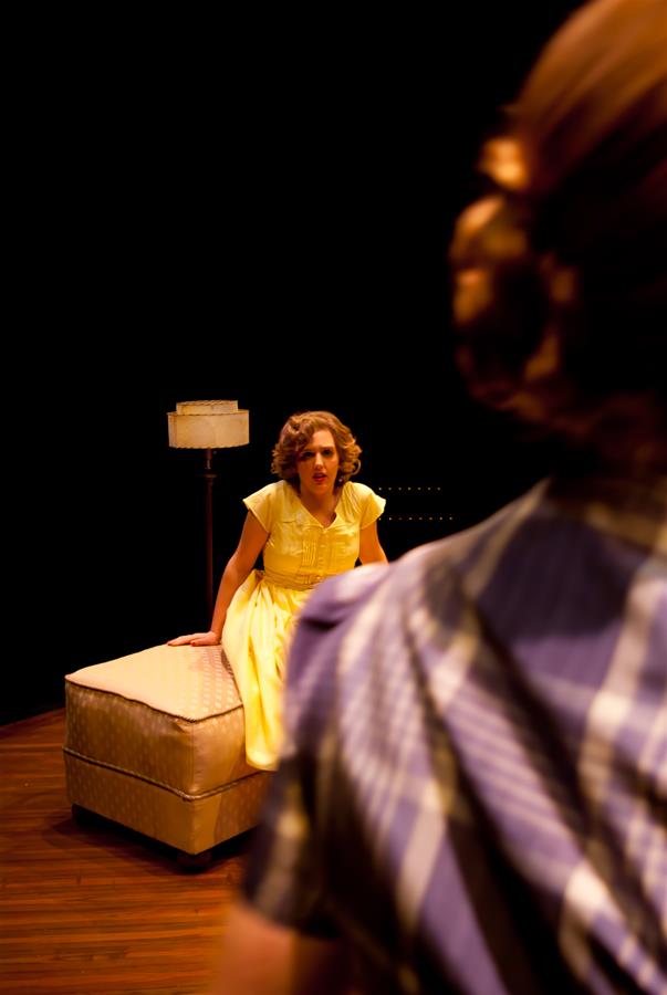 A woman in a yellow dress sits on an ottoman under a lamp on a stage. She appears to be speaking passionately. The foreground shows the back of another person's head and shoulder, slightly out of focus.