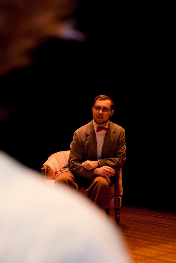 A man in a suit sits on a pink armchair on a dimly lit stage. He has glasses and a mustache. The focus is on him, with a blurred figure in the foreground. The background is dark, emphasizing the dramatic setting.