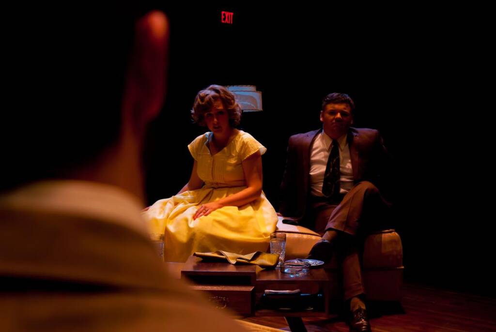 Two people sit on a couch on a dimly lit stage. The woman wears a yellow dress; the man is in a dark suit. Their expressions seem engaged with someone in the foreground. A red exit sign is visible in the background.