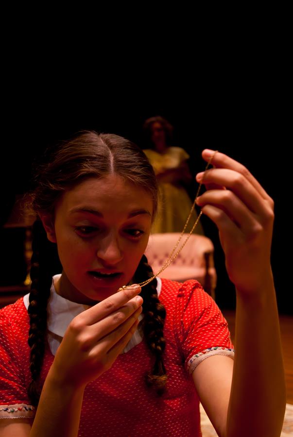 A young woman with braided hair, wearing a red dress with white details, intently examines a necklace. The background is dimly lit, with a person in a yellow dress and a chair barely visible.