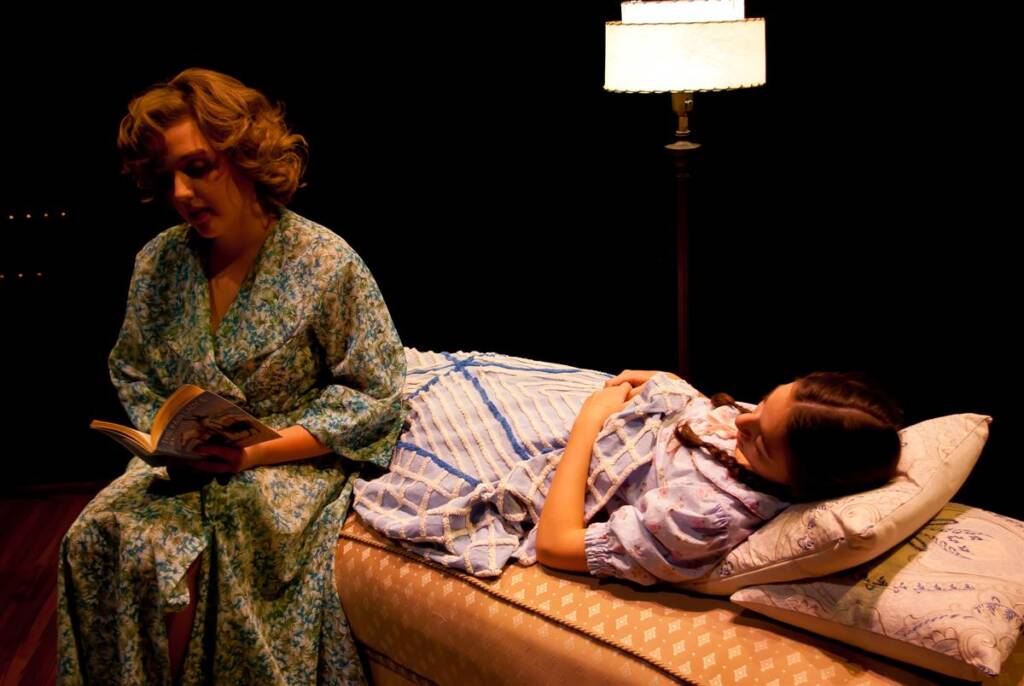 Two women in vintage dresses are in a dark room. One is sitting on the edge of a bed reading a book, while the other lies on the bed listening. A lit standing lamp provides soft illumination.