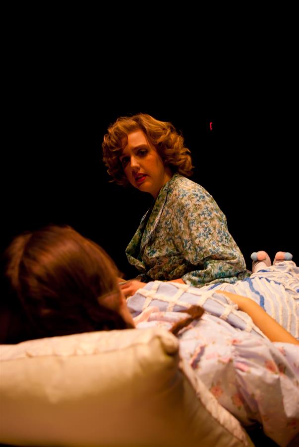 A woman with curly hair in a patterned robe sits beside a person lying on a bed. The lighting is dark and dramatic, highlighting their expressions in an intimate setting.