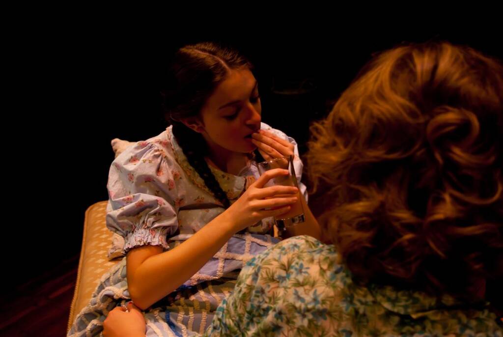 Two women sit facing each other in a dimly lit setting. One woman, wearing a patterned dress, drinks from a glass while holding her hand to her mouth. The other woman, with curly hair, has her back to the camera. The atmosphere is intimate and theatrical.